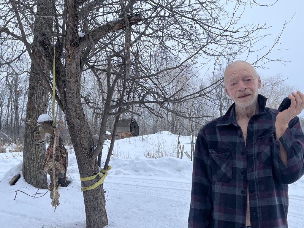Bald man with light skin and white beard wears plaid shirt and looks at camera, in front of snow-covered yard with deer carcass hanging from tree behind him.