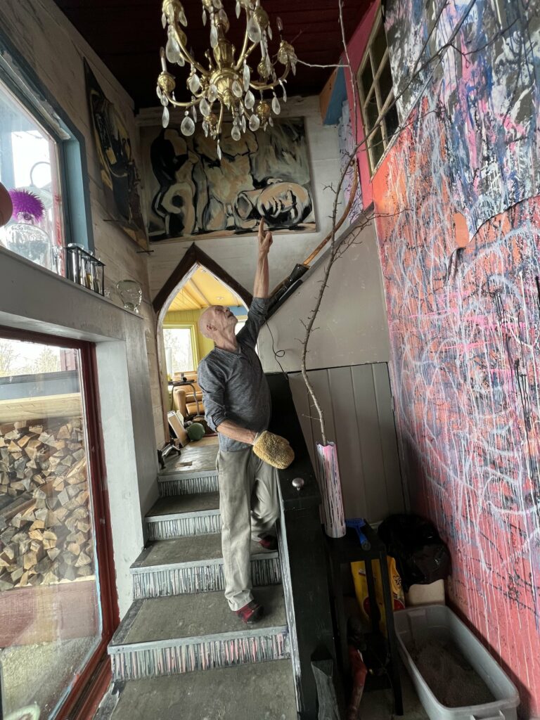 Person stands on staircase and points up at pink abstract painting, with large chandelier above and stacked wood visible through window.