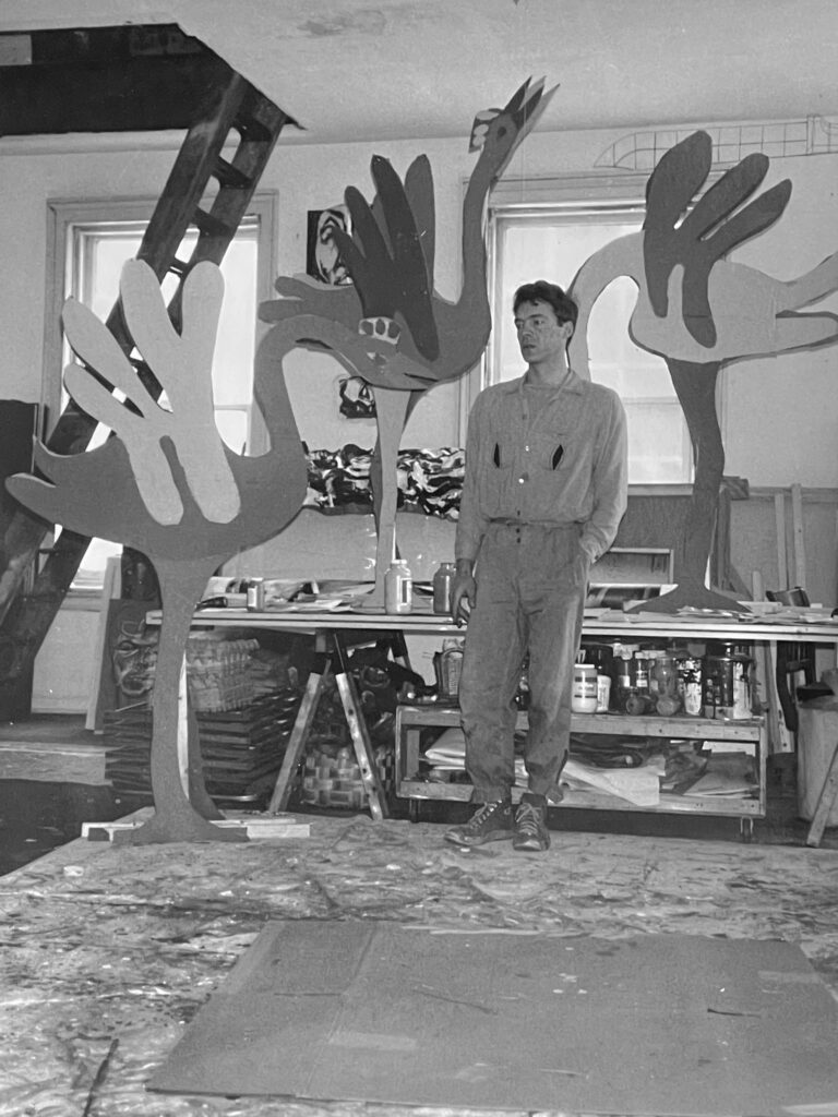Photo of artist in studio, with dropcloth on floor and large bird sculptures on table behind him.