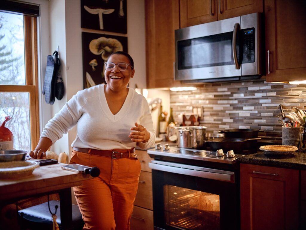 Person with medium dark skin, white sweater, orange pants, and glasses places hand on kitchen counter and leans forward laughing.