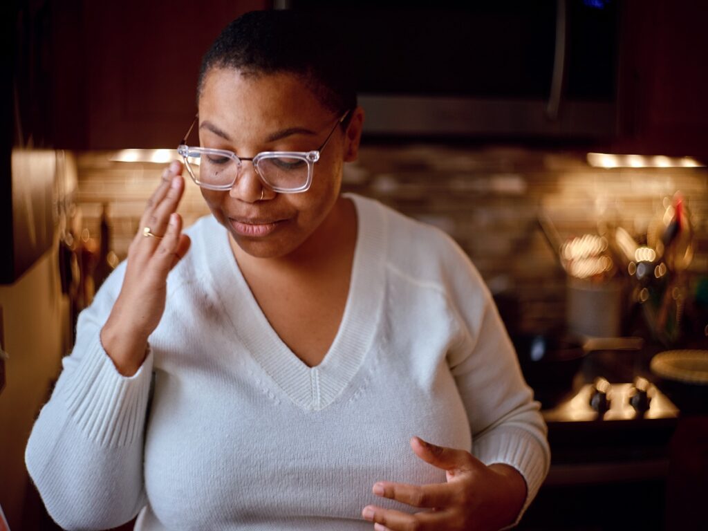 Person with medium dark skin, white sweater looks down slightly and raises hand to touch glasses.