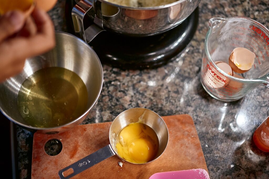 Metal mixing bowl, cup measure with egg yolks, and glass measuring cup with egg shells on kitchen counter.