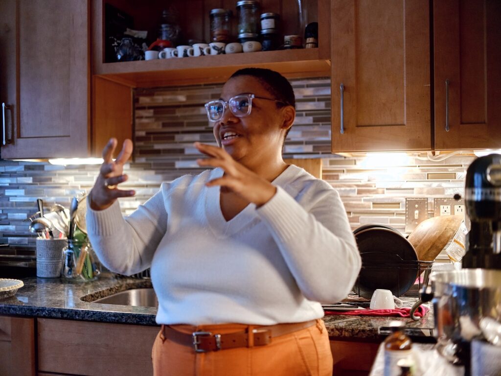 Person with medium dark skin, white sweater, orange pants, and glasses stands in front of stove and gestures outward with both hands.