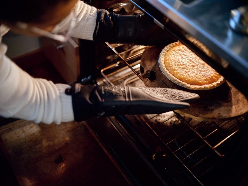 Hands in oven mitts reach for custardy yellow pie in oven.