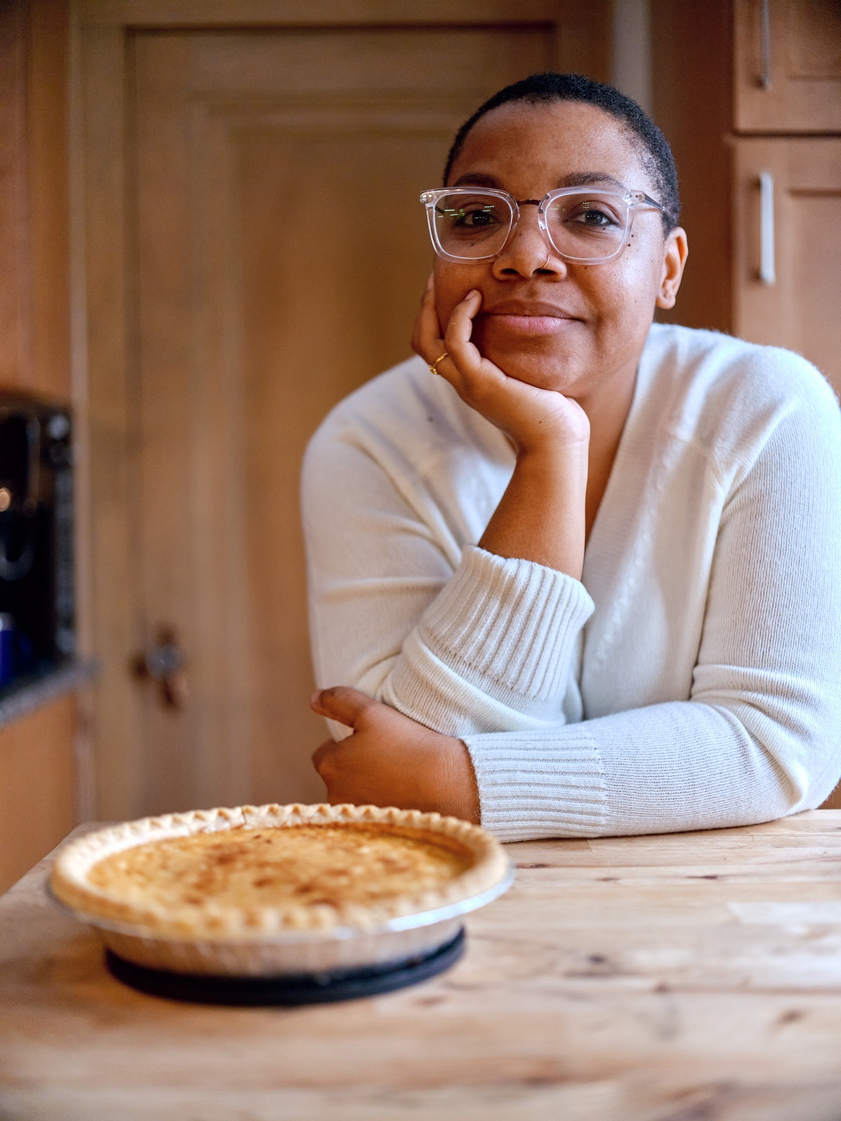 Person with medium skin, white sweater, and glasses leans on kitchen island resting hand on chin, with pie in front of them.