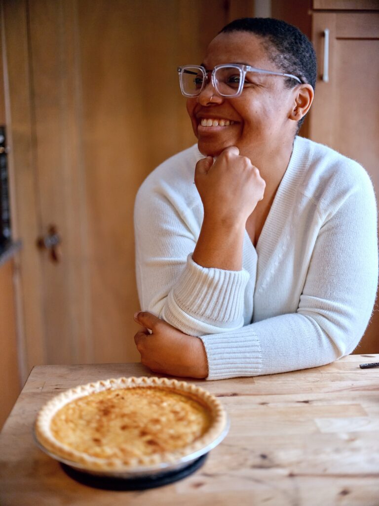 Person with medium skin, white sweater, and glasses leans on kitchen island in front of pie and looks to the side smiling.