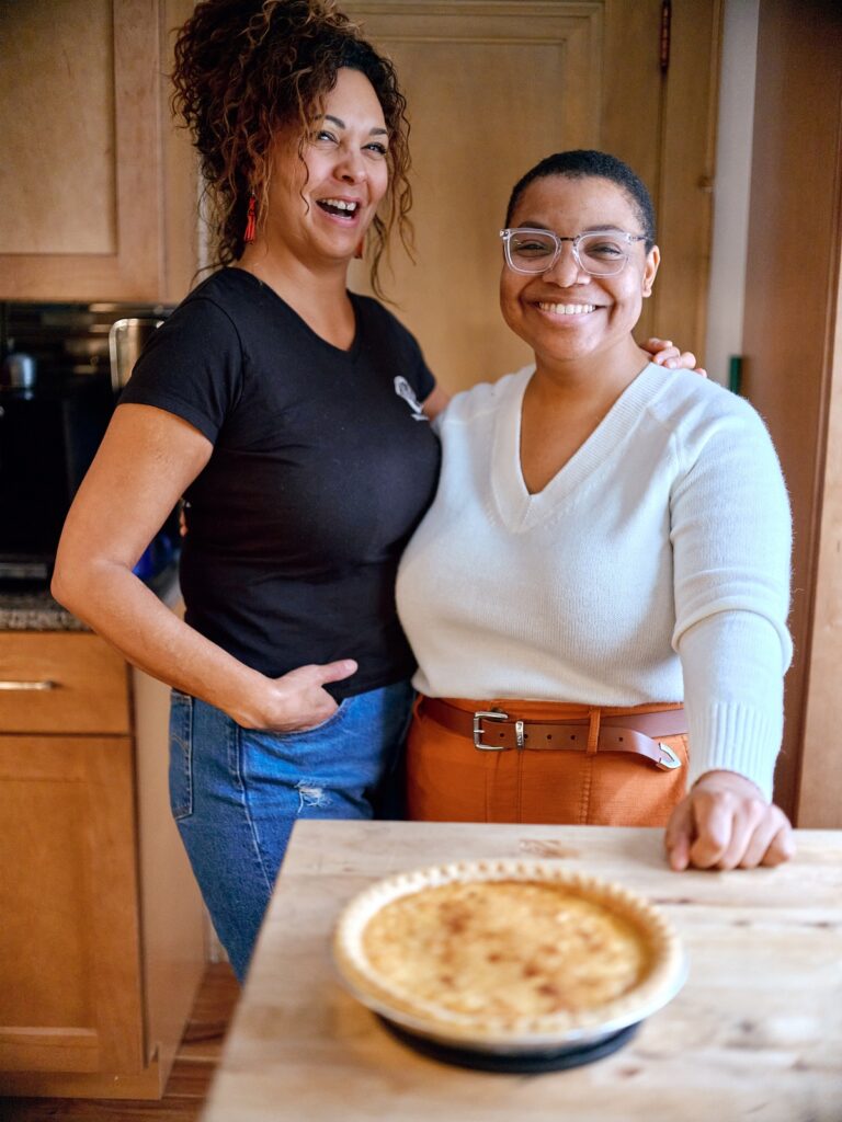 Two people stand together in front of pie on kitchen island, arms around each other and smiling.