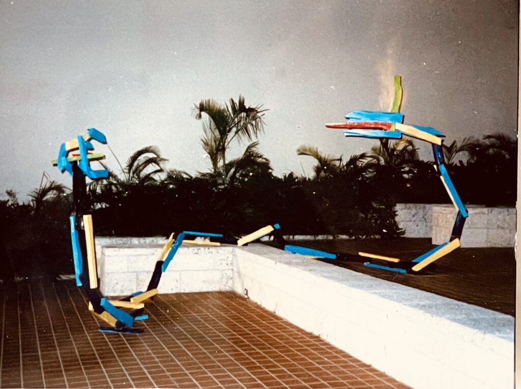 Abstract sculpture in blue, yellow, red, and black crosses over low white wall, with palm trees in background.