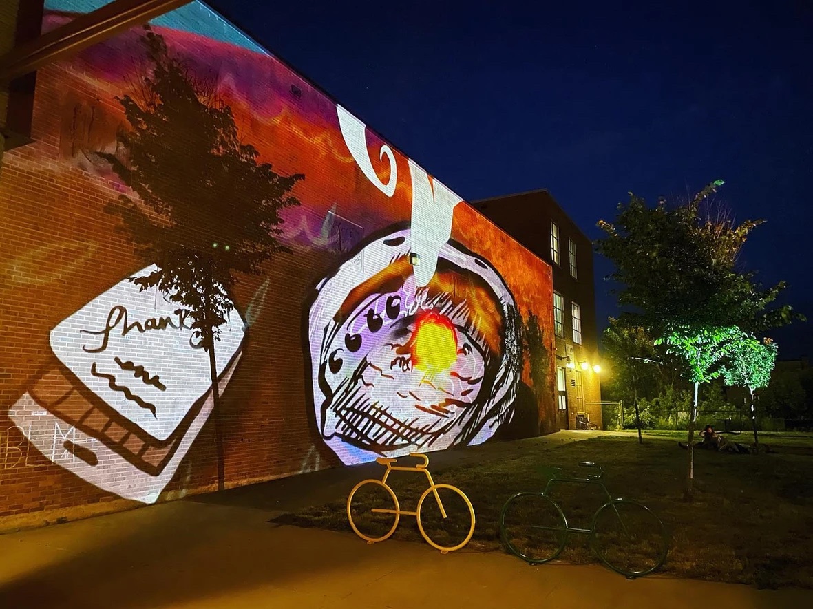 Large colorful projection on side of building at night, with bike racks and tree.