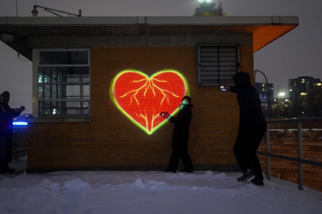 Three people stand outside small building at night, with projection of red and yellow heart on brick wall and snow on ground.