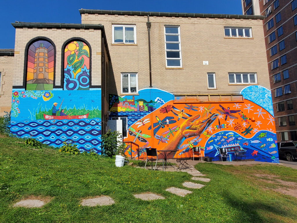 Mural on side of brick building, with person in red canoe on blue water and orange hand surrounded by colorful dragonflies.