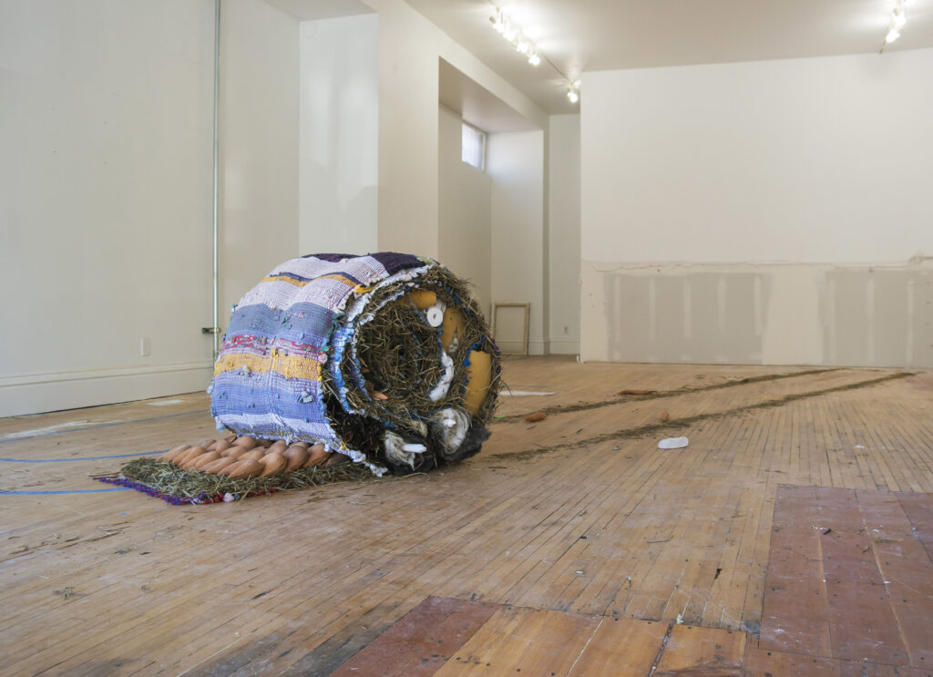 Large rolled sculpture in gallery with hay and braided rag rug.
