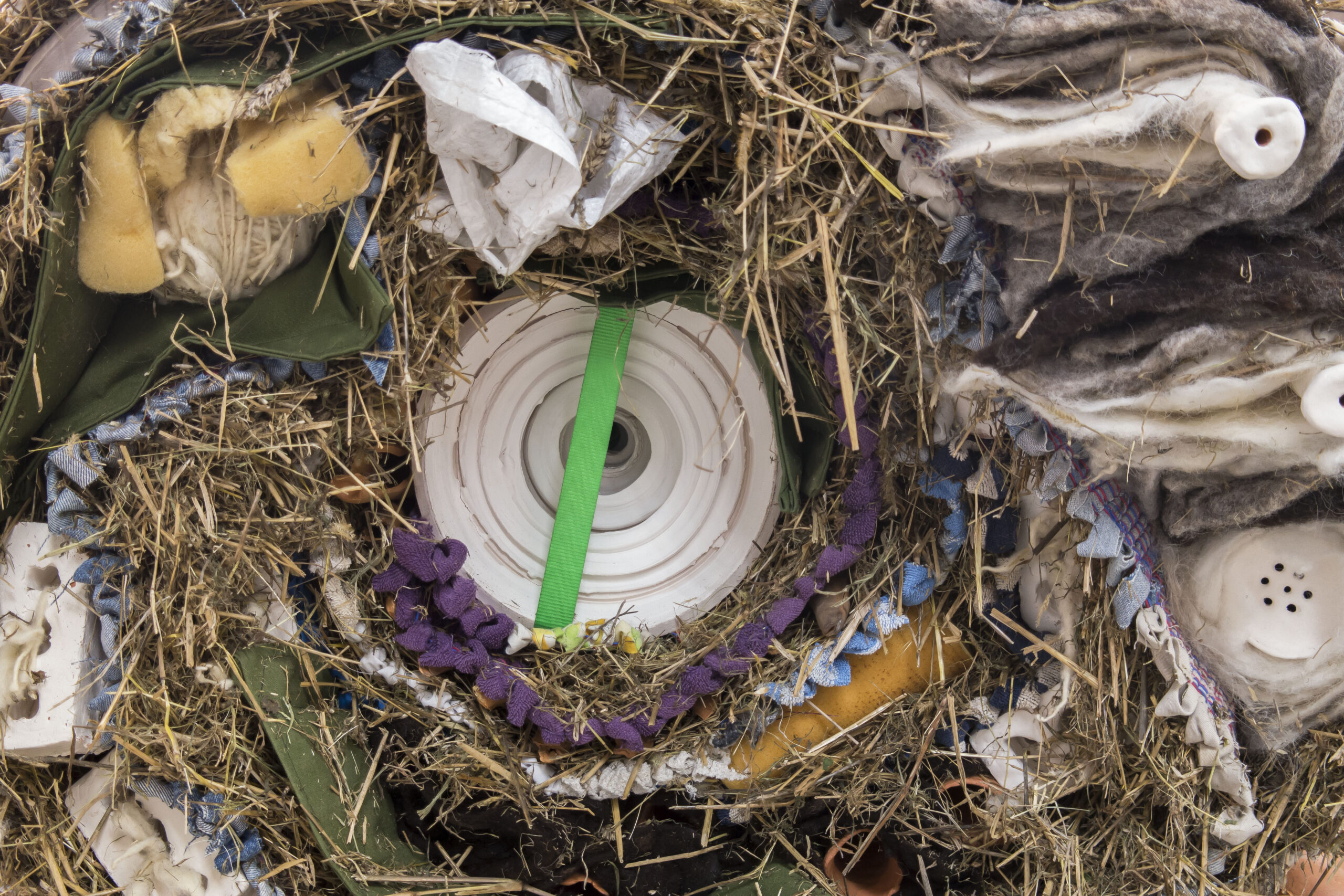 Detail photo of sculpture, rolled object with hay, braided rag rug, plastic and foam pieces.