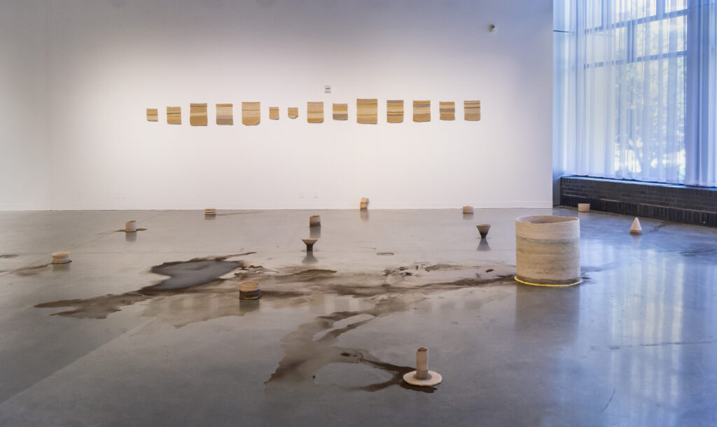 Gallery installation with small beige striped pieces on white wall, many small sculptures and dried brown liquid on floor.
