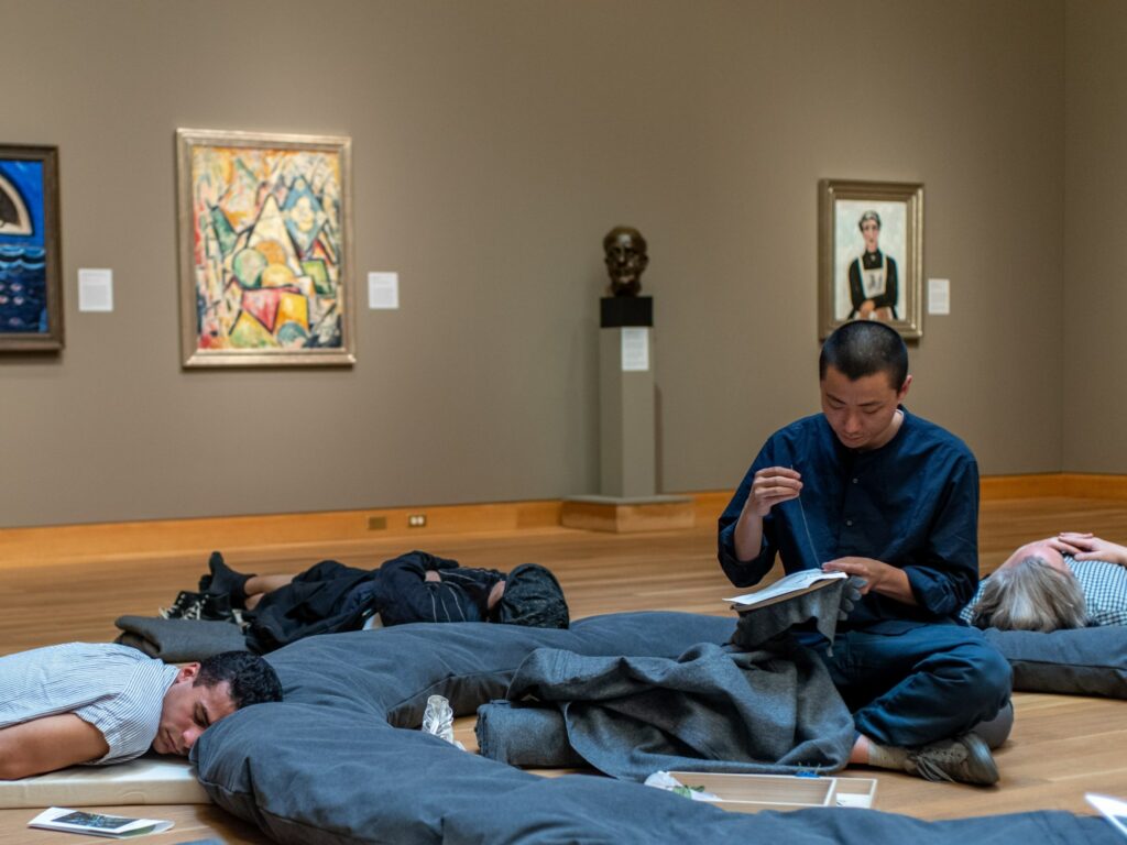 Artist dressed in blue embroidering, while people nap on gallery floor around him.