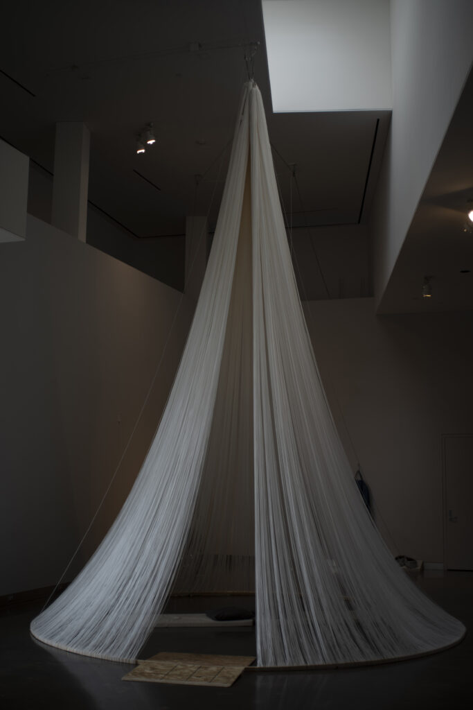 Sculpture installed in gallery, made of white strings in conical shape, with opening.