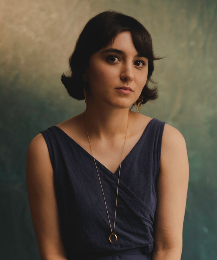 Person with light skin, brown bob, dark blue dress, and long gold necklace looks at camera.