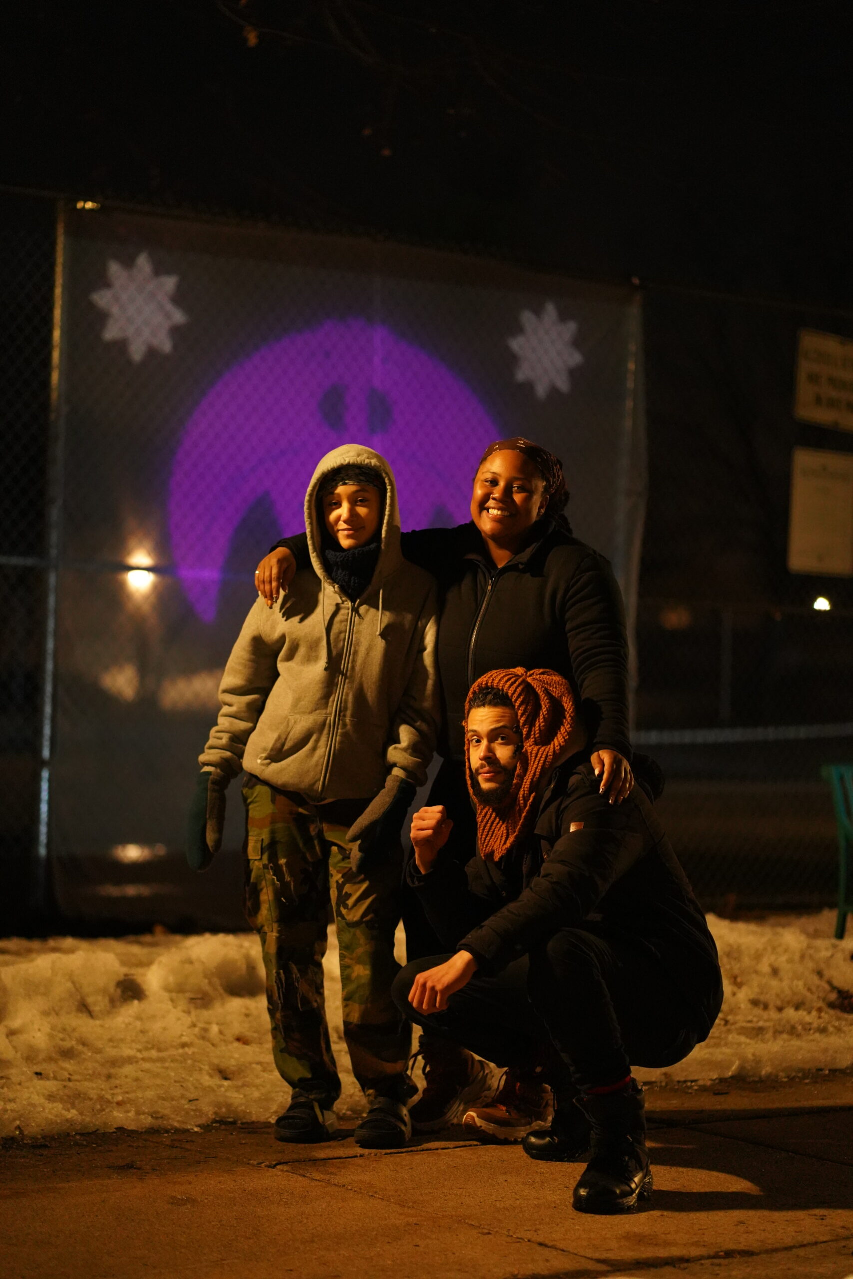 Three people in warm clothing stand with their arms around each other in front of projection on chain link fence.