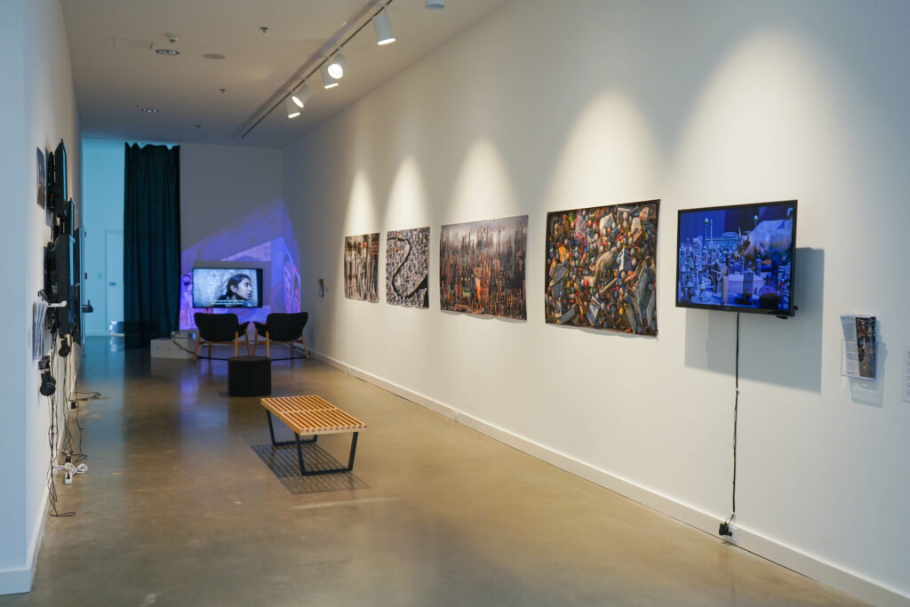 Long narrow gallery with video monitors, 2D works on walls, and benches and chairs.