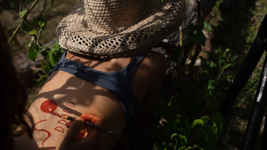 Person wearing straw hat lays on green foliage with back exposed, and someone else painting in red on the small of their back.