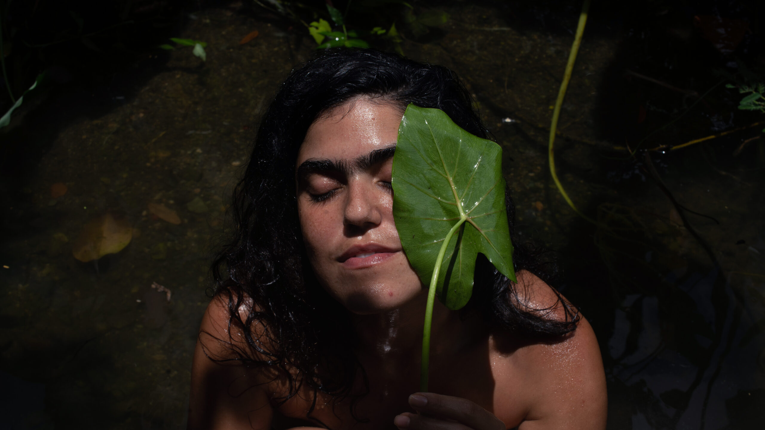 Person with long dark hair closes their eyes with sun on their face, behind thick green leaf.