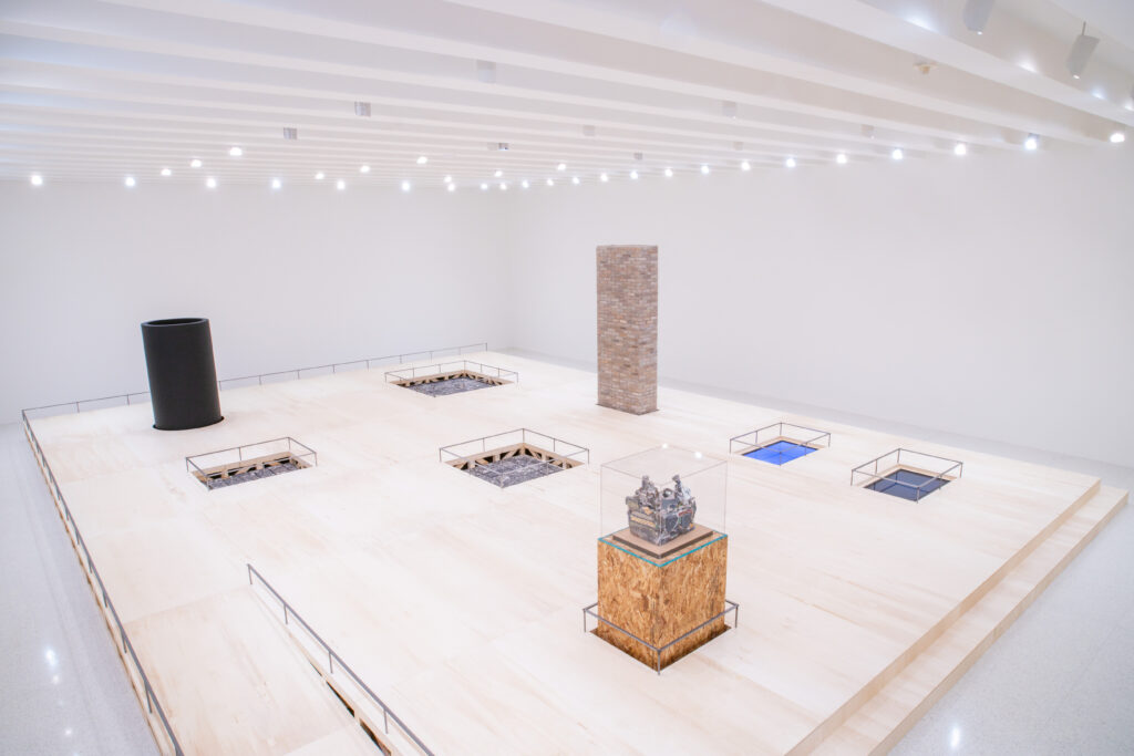 Angular view of gallery with white walls, white ceiling, and light wood flooring with several sculptures, some recessed into the floor.