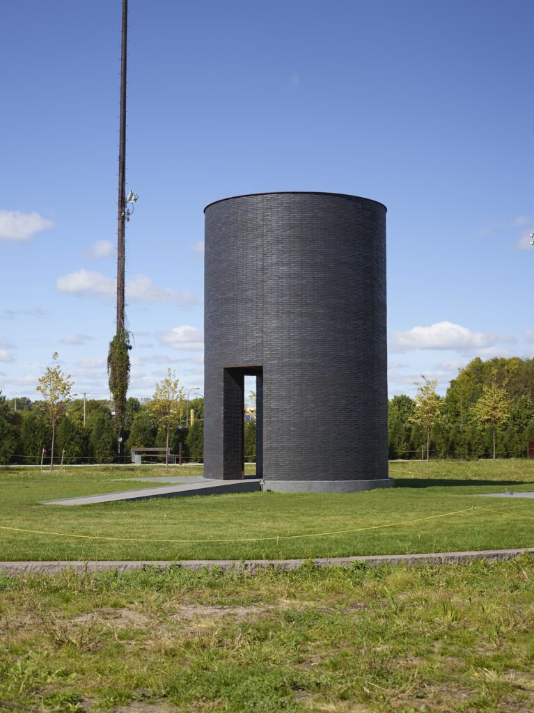 Large outdoor sculpture of black brick cylinder with ramp and open doorway, in green field with trees and blue sky behind.