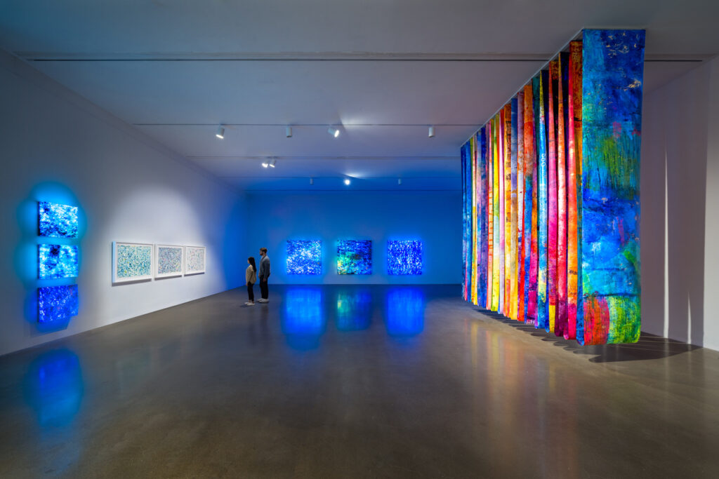 Gallery with artworks on wall, blue lightbox paintings on back wall, and row of large colorful panels hanging from ceiling.