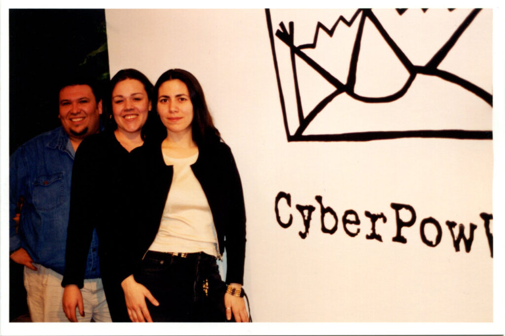 Three people stand close together smiling for camera in front of gallery wall with drawing and text reading "CyberPowWow".