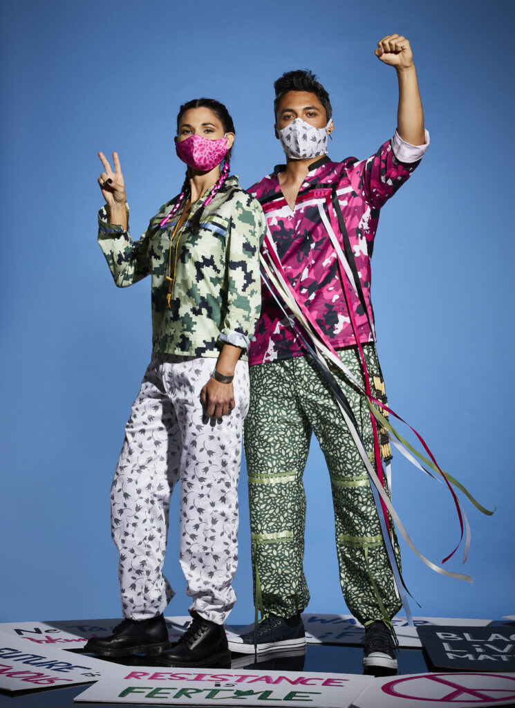 Two people wearing face masks, green and pink printed outfits with ribbons, with one holding fist and the other giving peace sign.