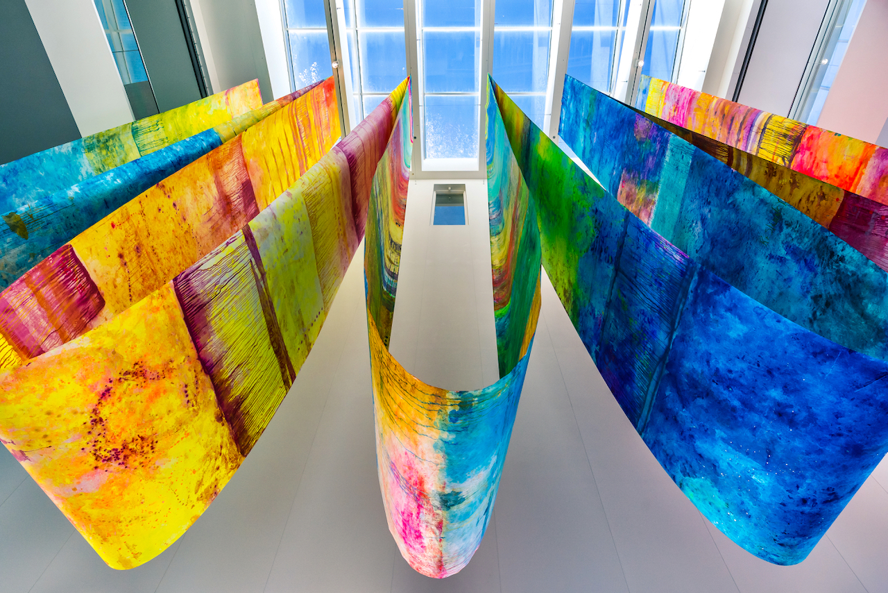 Large colorful fabrics draped from atrium ceiling, viewed from below with skylight above.