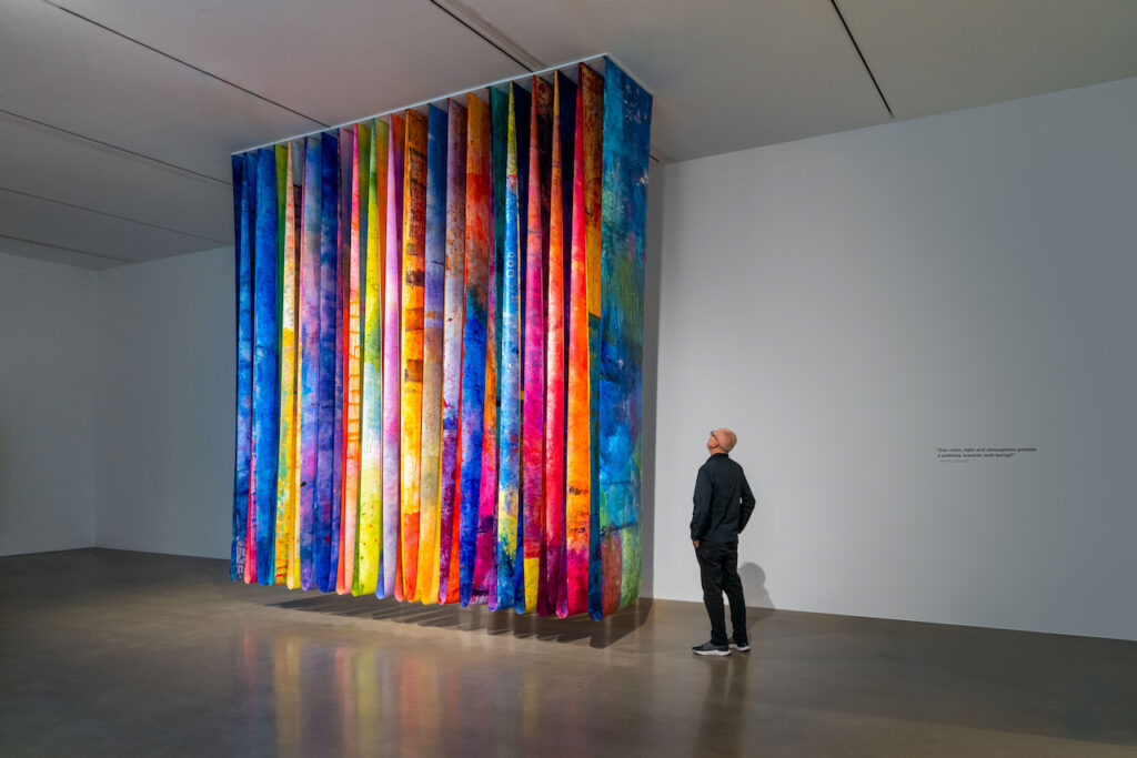 Person stands in gallery looking at row of large colorful panels hanging from ceiling.