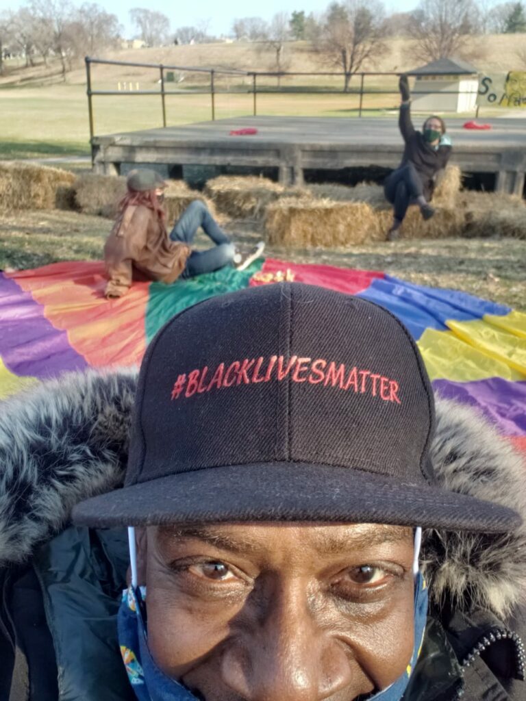 Person with dark skin, face mask pulled down, and Black Lives Matter hat takes a selfie with people, colorful blanket, hay bales, and outdoor stage in background.
