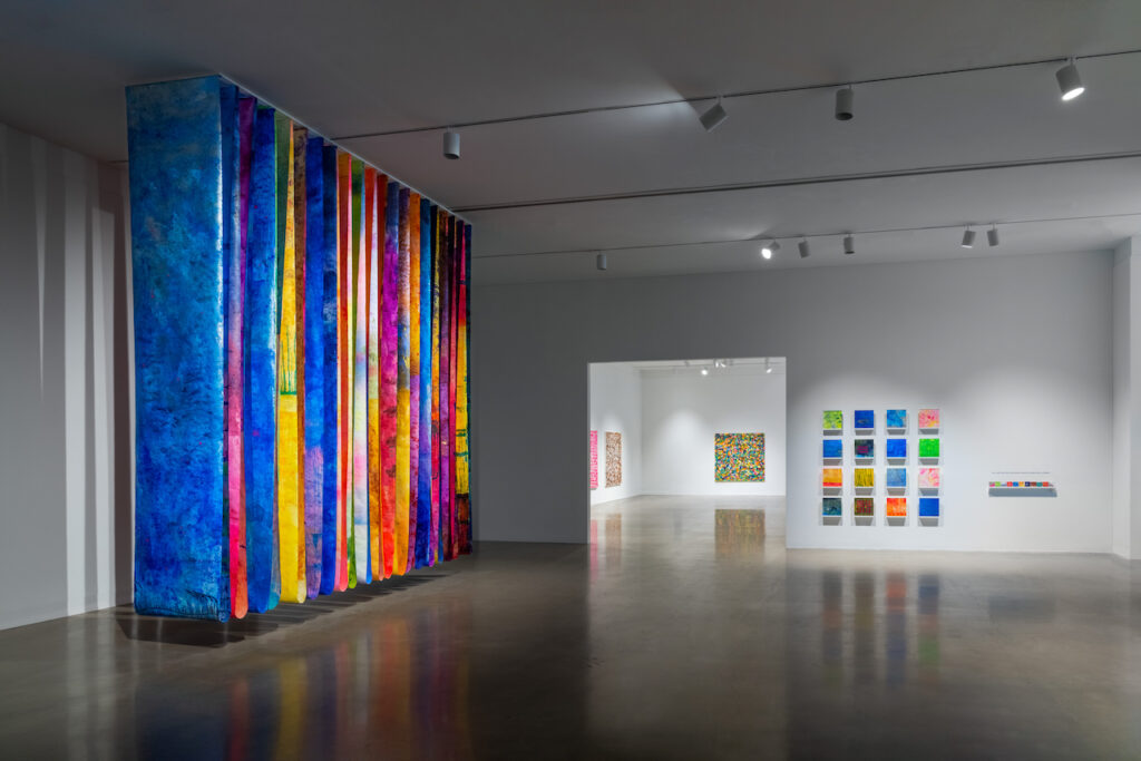 Gallery with row of large colorful panels hanging from ceiling, open doorway, and smaller colorful works on wall.