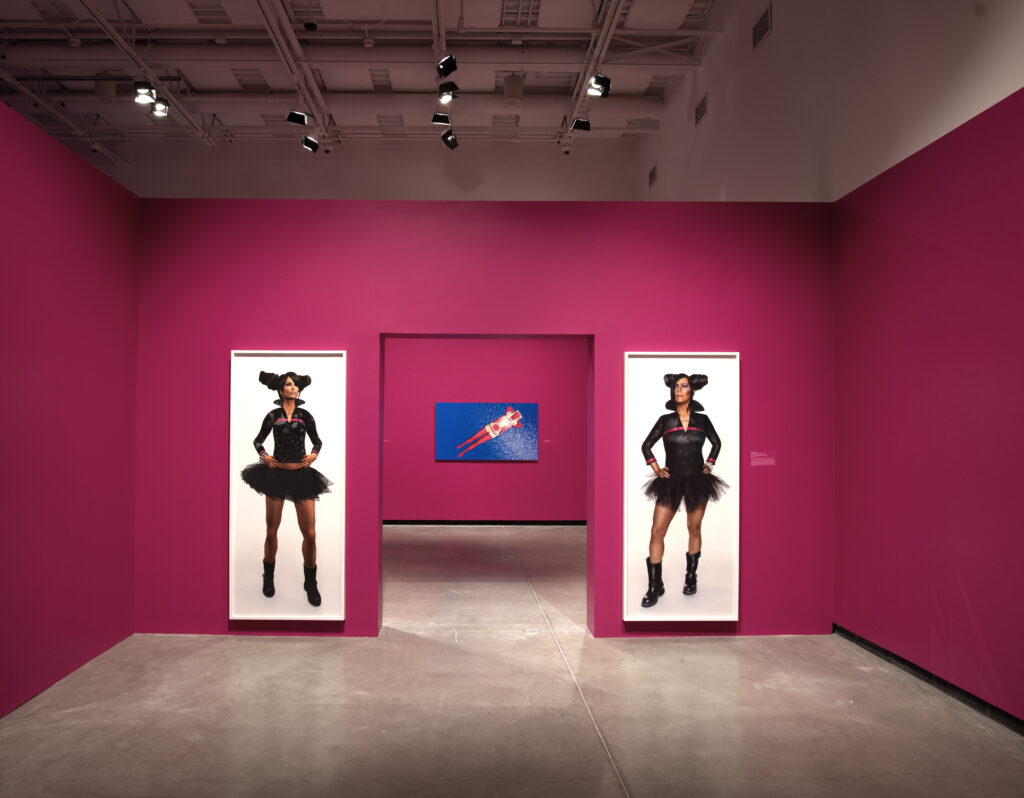 Gallery with bright pink walls, large images of two figures on either side of a doorway, and a blue 2D artwork on back wall.