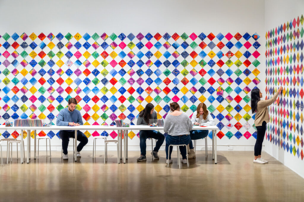 Colorful painted squares in diamond checker pattern fill two gallery walls, with people sitting at table.