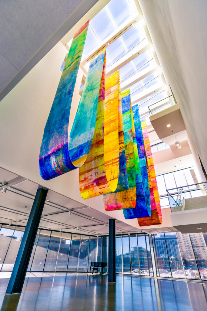 Large colorful fabrics draped from atrium ceiling, viewed from below with skylight above.