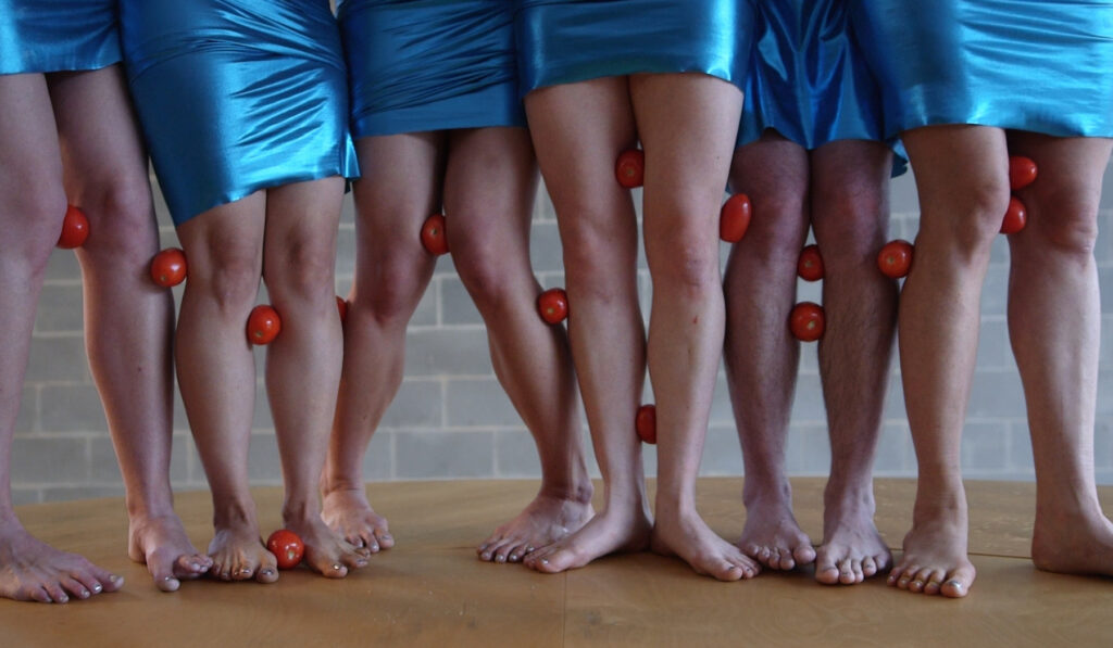 Six people wearing blue skirts stand close together with tomatoes held between their legs.