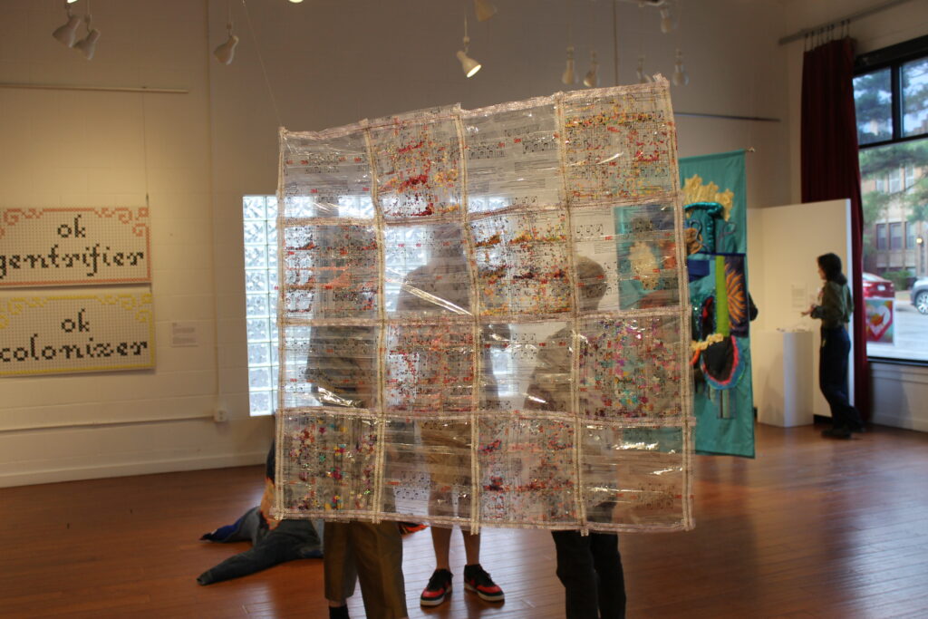 2D artwork of transparent material with square pattern and multicolored details hangs in middle of gallery.