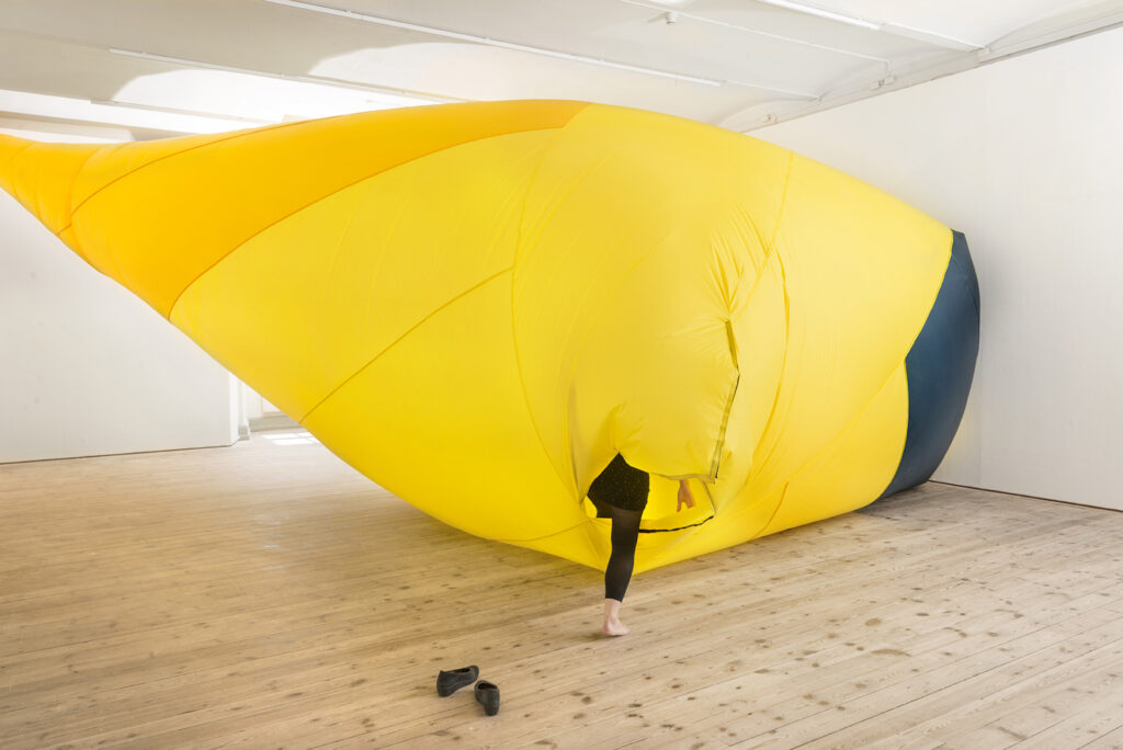 Large, bright yellow inflated object in gallery with leg of person stepping out of it.