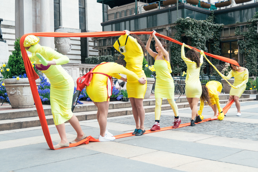 Seven performers wearing yellow stand in a line holding an orange band wrapped above and below the group.