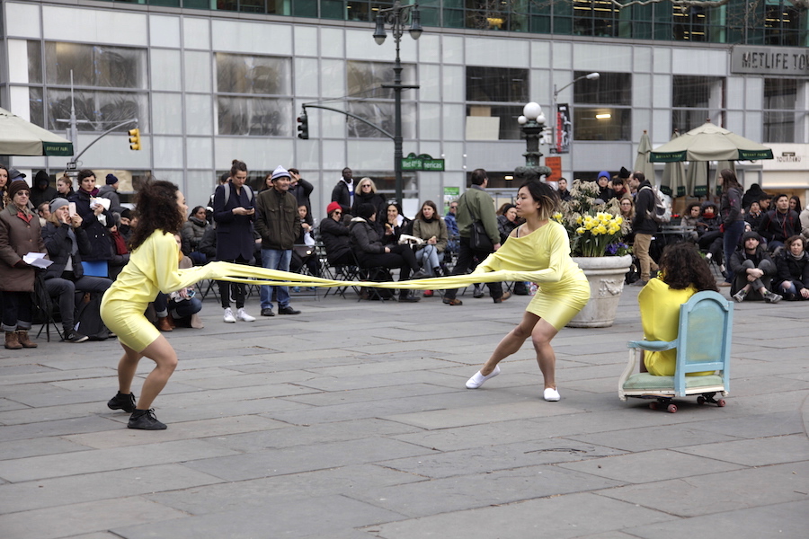 Three performers dressed in yellow perform in public square, one sitting in blue chair and the others wearing dresses attached to each other by long sleeves.