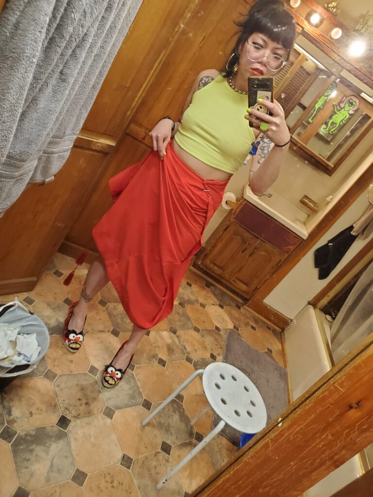 Person wearing orange skirt, neon yellow tank top, silver hoop earrings, and high heels holds a camera phone up to bathroom mirror.