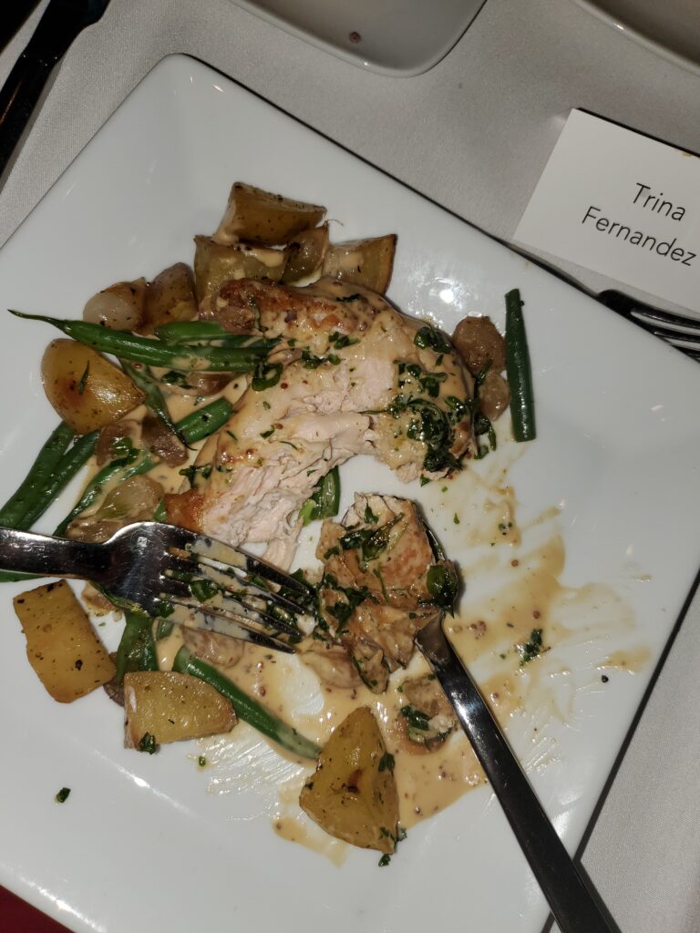 Square white plate with half-eaten chicken, potatoes, and green beans, with fork and spoon and a nametag reading "Trina Fernandez".
