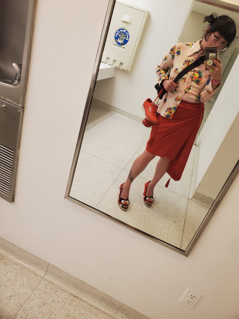 Person wearing red skirt, floral blouse, and heels holds phone up to bathroom mirror.