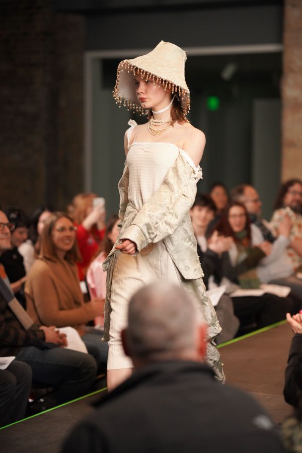 Person walks down runway wearing off-white long dress and hat with beaded lampshade.