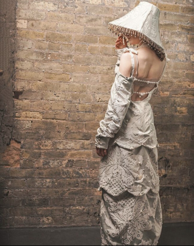 Person stands in front of brick wall with back to camera, wearing off-white long dress and hat with beaded lampshade.