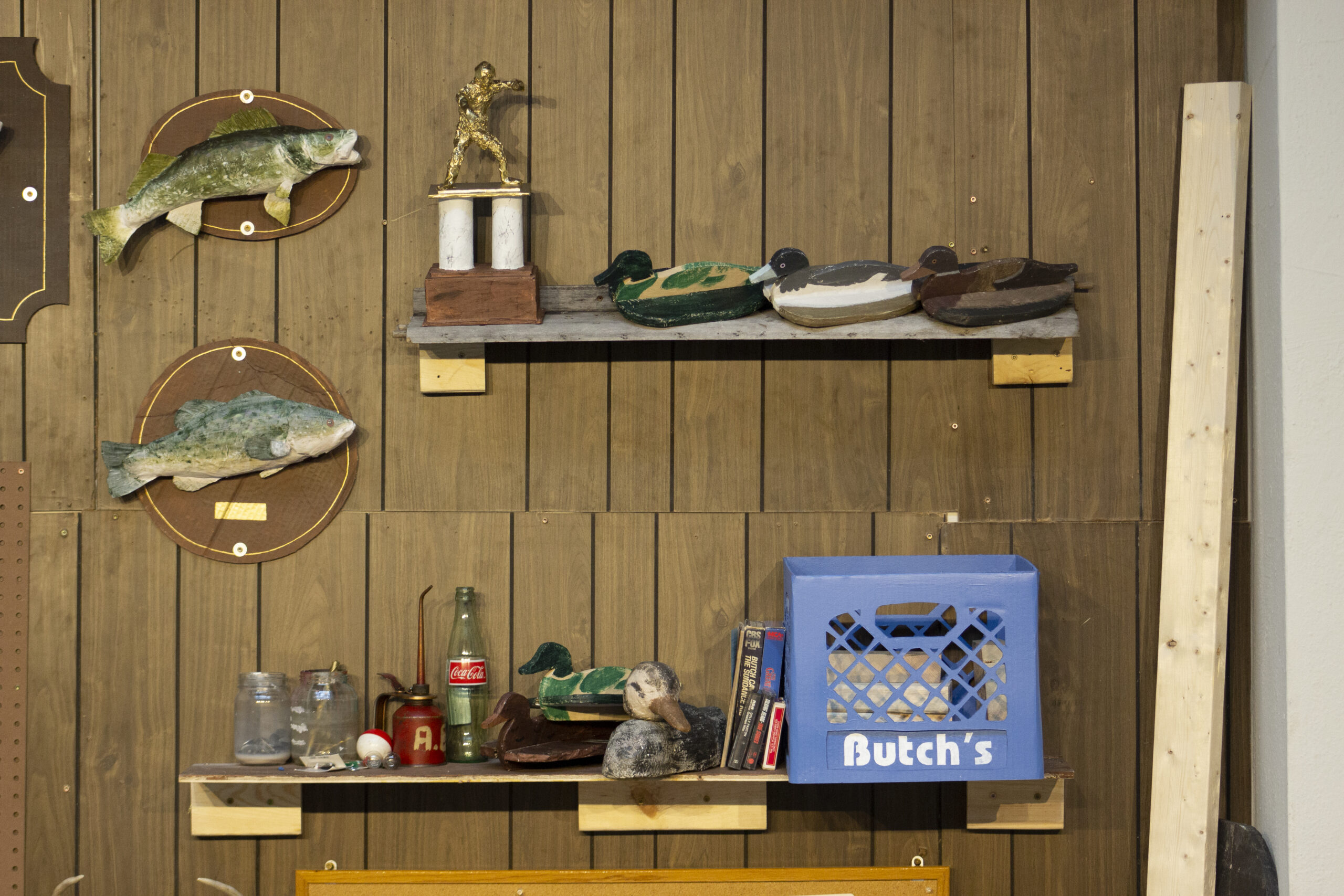 Installation on wood paneling with two mounted fish and two shelves with duck decoys, gold trophy, bottles, cassette tapes, and blue milk crate that says "Butch's."