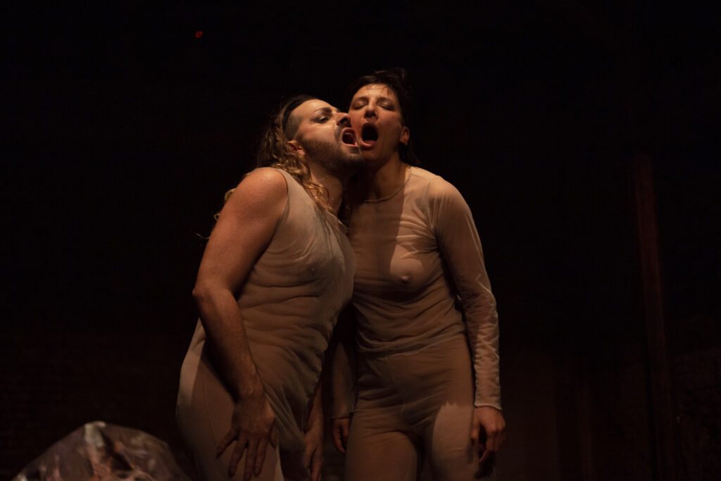 Two performers wearing tan clothing put their heads together and sing with open mouths.