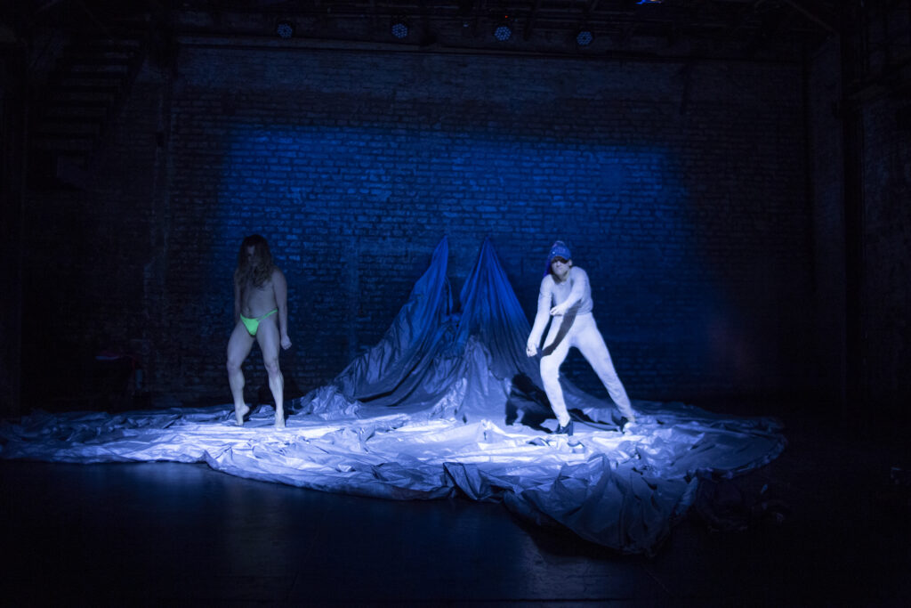 Two dancers step towards the audience on large fabric, bathed in blue light.